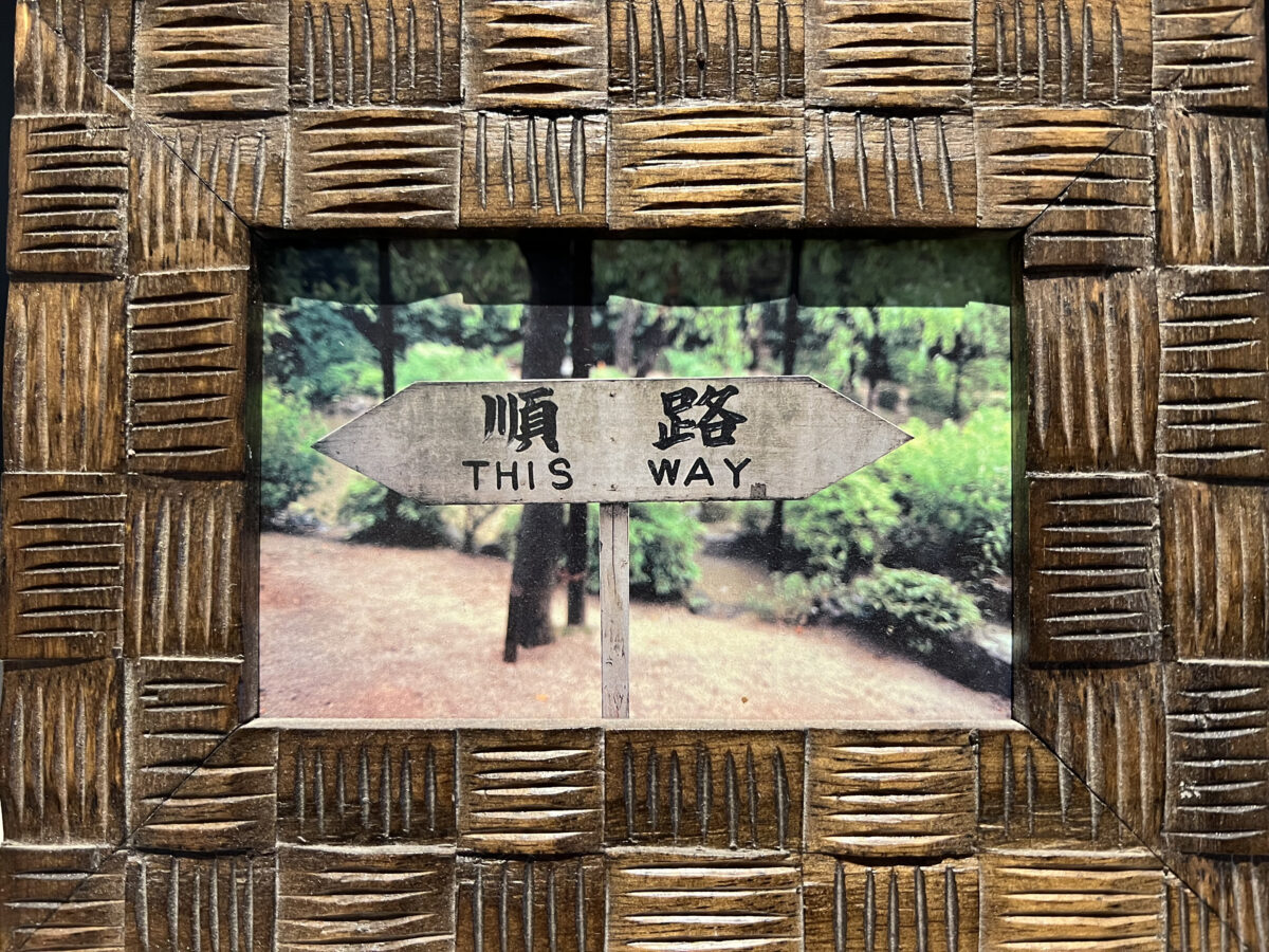 A tactile earthy wooden frame with a photo in it with a "This Way" sign written in Chinese and English, in the forest with a trail in the background